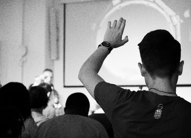 Kopa ar cilvēkiem, priekšplānā vīrietis ar paceltu roku jautājuma uzdošanai. Foto: unsplash.com